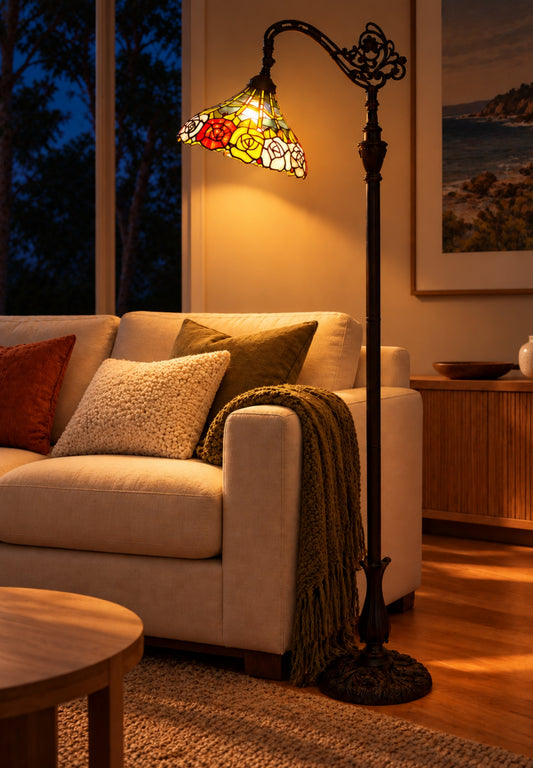 A classic black metal floor lamp with an ornate scrollwork top and a colorful stained-glass shade featuring red, yellow, and green floral motifs, shown lit in a cozy living room next to a beige sofa.